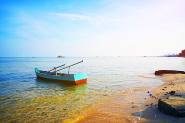 Naklejka premium Boat on shore at beach during late afternoon with clear sky and calm water in background. Small Fishing boat sits near shoreline on sea sandy beach. Ocean is clear and calm. Fishing wooden boat in sea
