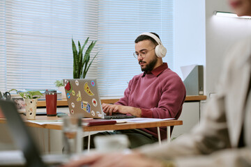 Young adult Middle Eastern man wearing headphones working on laptop at desk in modern office,...