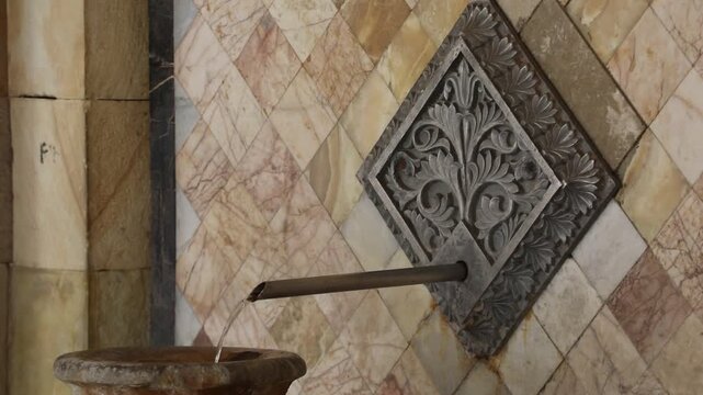 Ornate metal spout pours mineral water into a stone vase against a textured marble tile wall in a historic spa setting, showcasing natural thermal spring source details.

