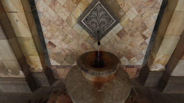 Ornate metal spout pours mineral water into a stone vase against a textured marble tile wall in a historic spa setting, showcasing natural thermal spring source details.

