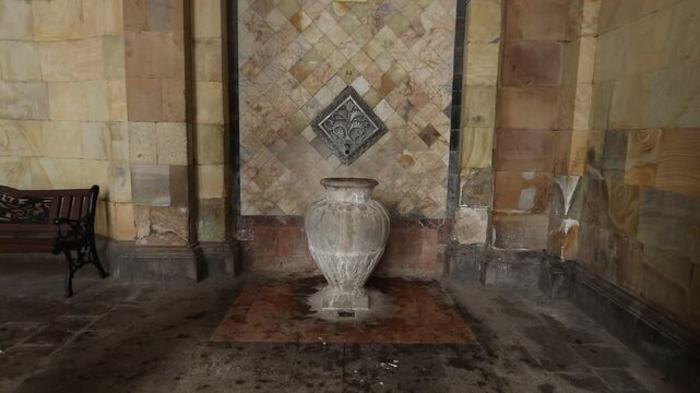 Ornate metal spout pours mineral water into a stone vase against a textured marble tile wall in a historic spa setting, showcasing natural thermal spring source details.

