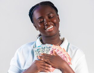 A smiling young woman with braided hair joyfully hugs a fan of ghana cedi banknotes to her chest. She wears a white polo shirt, her eyes closed in a moment of pure financial bliss and contentment.
