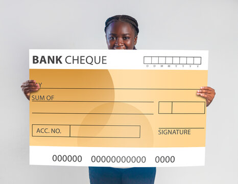 An African woman smiles behind a large, oversized novelty bank check. The yellow and white ceremonial check features blank fields for a date, payee, sum, and signature against a clean grey backdrop.