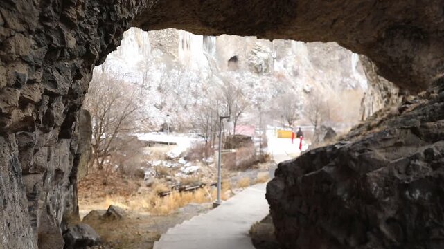 Paved hiking trail passes directly underneath massive natural stone archway inside deep rocky canyon with steep cliffs and patches of white snow during cold cloudy winter day outdoors.

