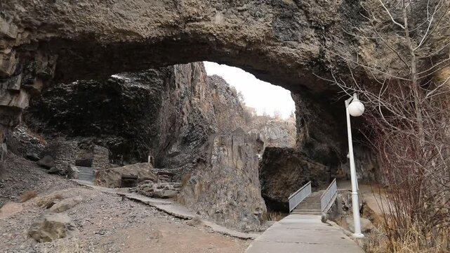 Paved hiking trail passes directly underneath massive natural stone archway inside deep rocky canyon with steep cliffs and patches of white snow during cold cloudy winter day outdoors.


