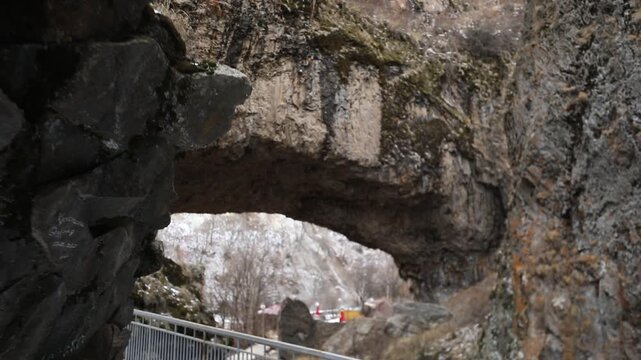 Paved hiking trail passes directly underneath massive natural stone archway inside deep rocky canyon with steep cliffs and patches of white snow during cold cloudy winter day outdoors.

