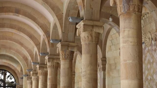 Tilt down view of a historic stone gallery interior featuring high arches, detailed columns, and a skylight ceiling, with pine trees visible through the large glass windows.

