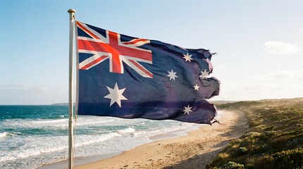 Frayed Australian national flag waving proudly above a rugged coastal beach landscape under a bright clear sunny sky during daytime hours