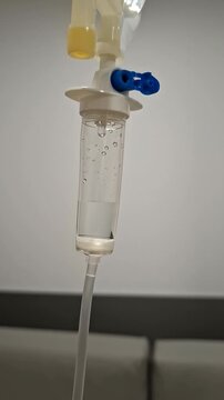 A drop of clear medicine drips steadily into an intravenous line. The setting is a hospital or clinic. This process is a part of patient treatment during recovery.