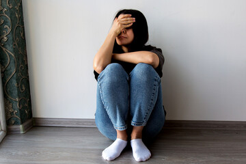 Sad young woman sitting on the floor with her knees drawn up, resting her head on her hand,...