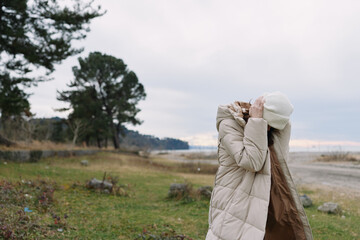 Obraz premium Outdoor woman in a beige winter coat and white hat adjusts her hood beside a foggy landscape with lake, trees and grass, conveying peaceful solitude and seasonal fashion in nature