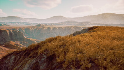 Obraz premium Vibrant grass sways gently atop a rocky ridge as golden light filters through the evening sky, illuminating rugged mountains and valleys in the distance, creating a serene atmosphere of nature.