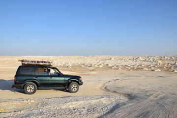 Off-road vehicle in the White Desert. Car driving across a vast Sahara landscape with unique chalk rock formations under a clear blue sky. © Aleksei