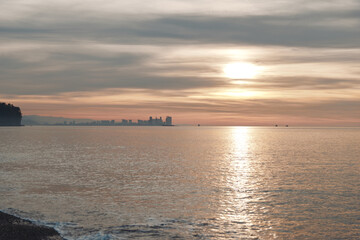 Fototapeta premium Sunset over calm water with a distant city skyline on the horizon, gentle reflections on the sea, and a quiet shore framing a serene evening seascape.