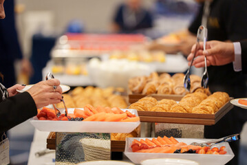 People serving themselves food with tongs from plates at a buffet table. Hands selecting a variety of dishes in a self-service dining setup, typical for events, parties, or restaurants