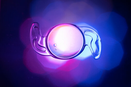 Macro Shot Of A Flexible Intraocular Lens (IOL) With Haptics On A Neon Blue And Purple Backgr