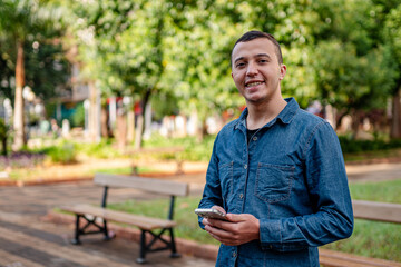 Fototapeta premium Happy young man in a blue denim shirt holding a smartphone and smiling while standing in a sunny outdoor park.