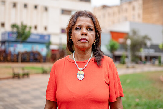 Portrait of a serious mature Black woman in an orange shirt standing in an urban park.