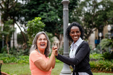 Fototapeta premium Two diverse senior women holding hands and laughing together in a city park, celebrating friendship and active aging.