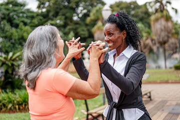 Naklejka premium Two diverse senior women holding hands and laughing together in a sunny outdoor park.