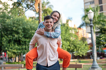 Obraz premium Happy young man giving his girlfriend a piggyback ride in a sunny city park, both smiling and looking at the camera during a romantic date outdoors.