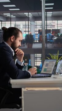 Vertical Video Professional man engaged in company data analysis with laptop and tablet, reviewing KPIs and profitability metrics for decision making in firm administration. Executive office tasks.