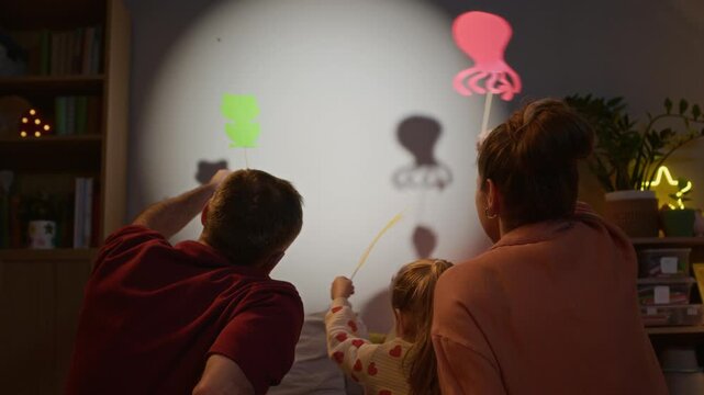 Back view shot of unrecognizable family sitting on bed, playing shadow puppets and creating store while sharing warm family moment