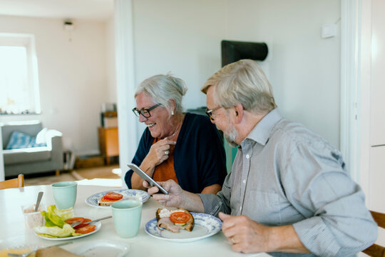 Senior couple laughing over smartphone at home kitchen table