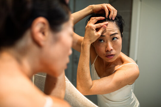 Woman inspecting scalp for hair loss in bathroom mirror