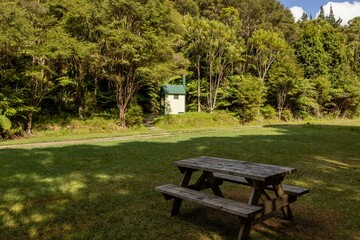 Fototapeta premium A picnic table sits on a grassy lawn in Broken Hills Valley, New Zealand. A small outhouse with a green roof is nestled in the trees. This is a popular spot for tourists to stop and camp.