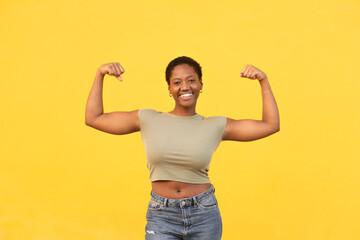 Fit healthy woman flexing muscles against yellow background. Smiling young black lady showing...