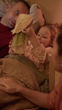 Vertical shot of little daughter lying in bed, playing with plush rabbit and chatting with her beloved parents
