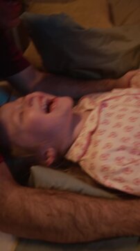 Vertical handheld shot of smiling parents playing with little daughter who lying on bed and laughing out loud