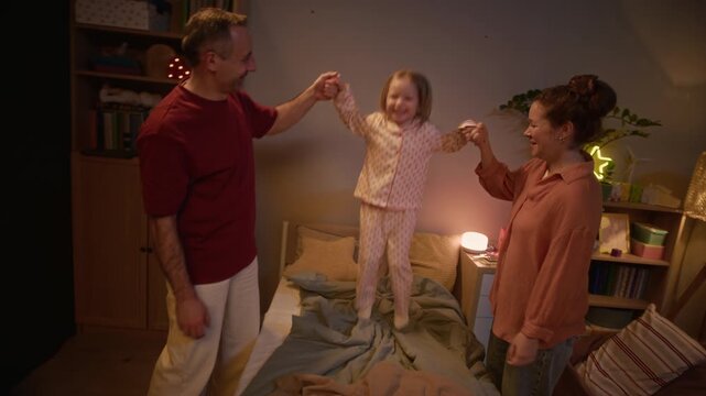 Slow motion of little blonde girl holding parents hands and jumping on bed, her mother and father admiring her