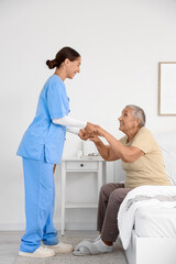 Female caregiver helping happy senior woman to stand up in bedroom