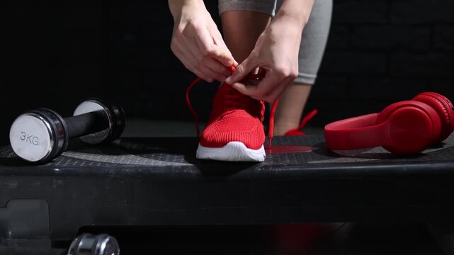 Woman tying shoelace of red sneaker near bottle, headphones and dumbbell against black background indoors, closeup. Camera moving left