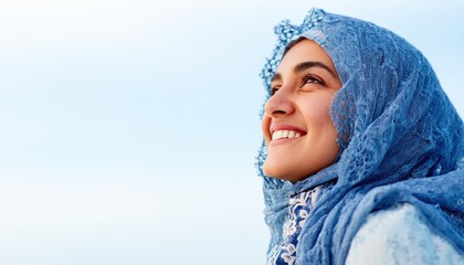 Young woman wearing a blue headscarf looks upward with a joyful expression against a bright sky.