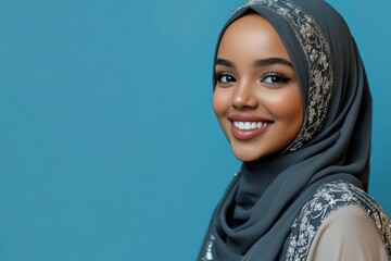 Young woman wearing an intricately decorated headscarf smiles brightly against a solid blue backdrop