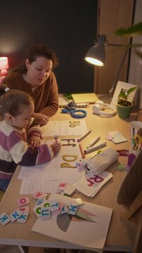 Vertical shot of brunette mother sitting near her daughter who writing letters on paper, improving handwriting skill