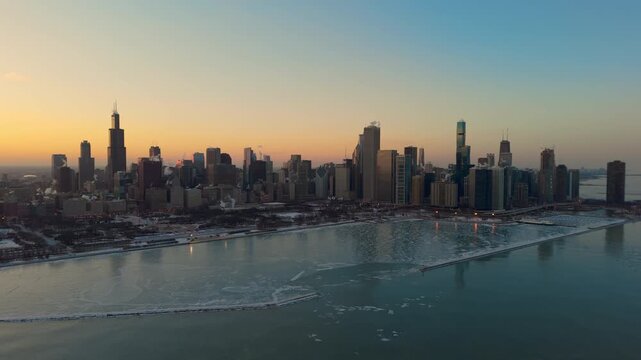 Drone view of downtown Chicago skyline at sunset during winter. Ice formations float on the surface of Lake Michigan as subzero temperatures create frozen patterns along the shoreline