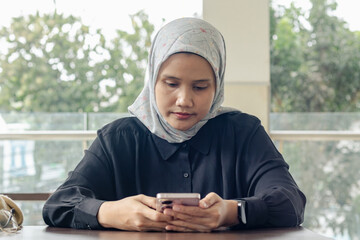 A young Indonesian Muslim woman sitting in a coffee shop while replying to messages on her smartphone