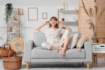 Obraz premium Young pretty woman in headphones sitting on sofa and listening music in light living room