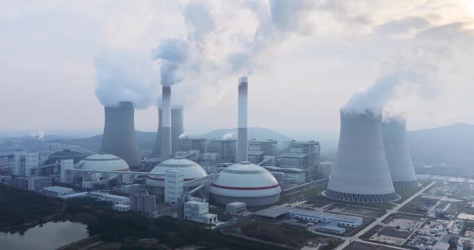 Aerial drone footage orbit around a coal-fired power plant at dawn: cooling towers, smokestacks emitting steam, and industrial buildings under soft morning light. 
