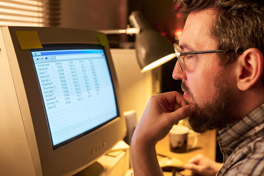 Caucasian man wearing glasses working at desk, analyzing spreadsheet data on computer monitor, resting chin on hand, focused on screen in office setting
