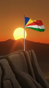 Seychelles flag flies on a high pole on the edge of high rock hill against sunset sky