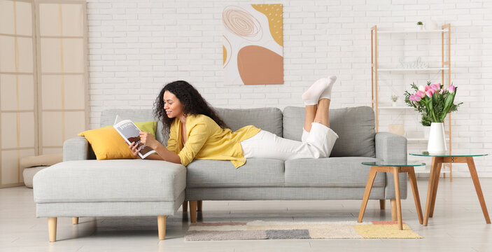 Young African-American woman with tulips reading magazine at home on spring day