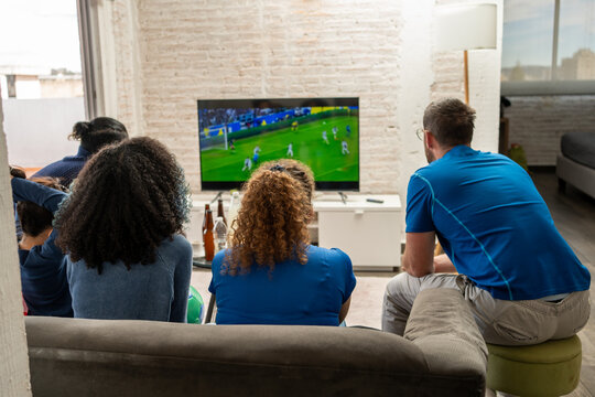 Family and friends seen from behind watching football soccer on television, the screen visible as the match occurs at home. Sports viewing, shared focus, friendship, togetherness, indoor gathering