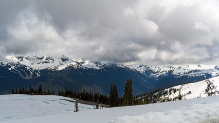 Fototapeta premium Whistler gondola crossing with views of alpine peaks, valley floor, and patchy snow under a cloudy sky.
