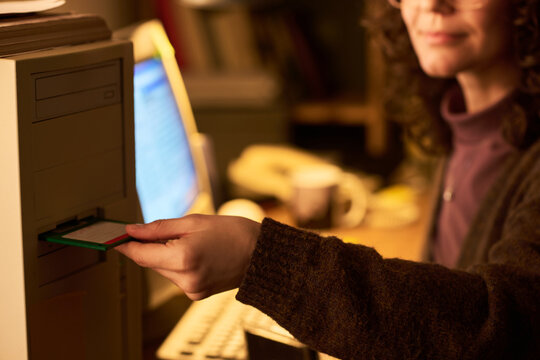 Caucasian woman inserting floppy disk into vintage desktop computer, sitting at desk with monitor in background, focusing on using outdated technology in office setting