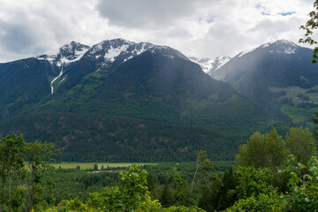 Fototapeta premium Snow-capped mountain peaks with forested slopes above flat river valley.